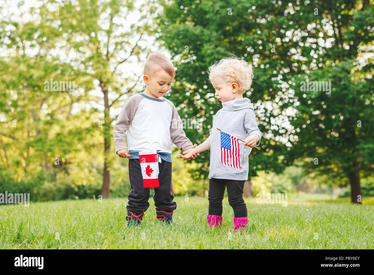 Heureux adorable petit blond Caucasian girl and boy smiling rire tenant la main et en agitant des drapeaux américains et canadiens, à l'extérieur dans le parc, la célébration de Banque D'Images