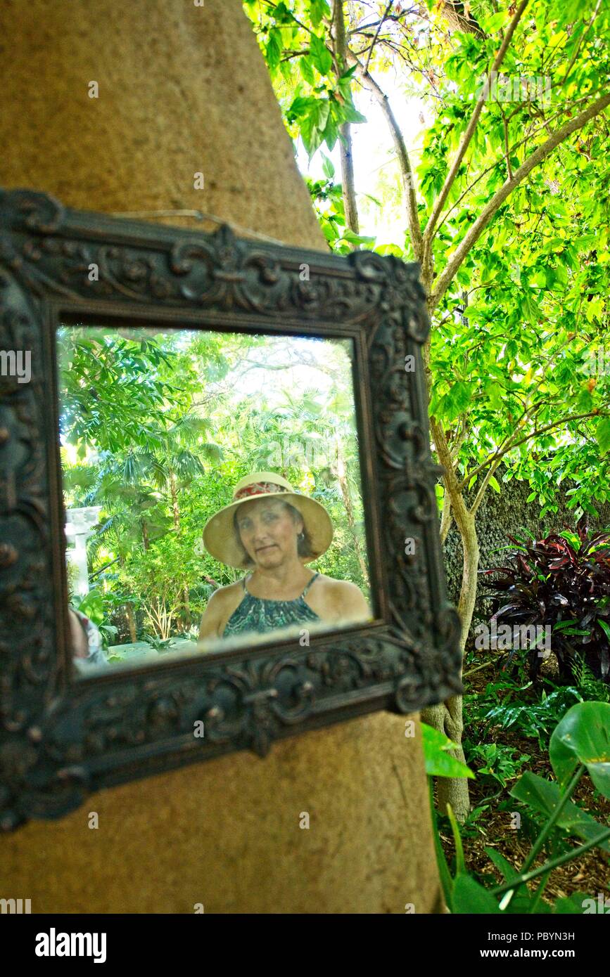 Le reflet d'une femme portant un chapeau de soleil et en robe d'un miroir suspendu à un arbre à l'extérieur en milieu tropical Banque D'Images