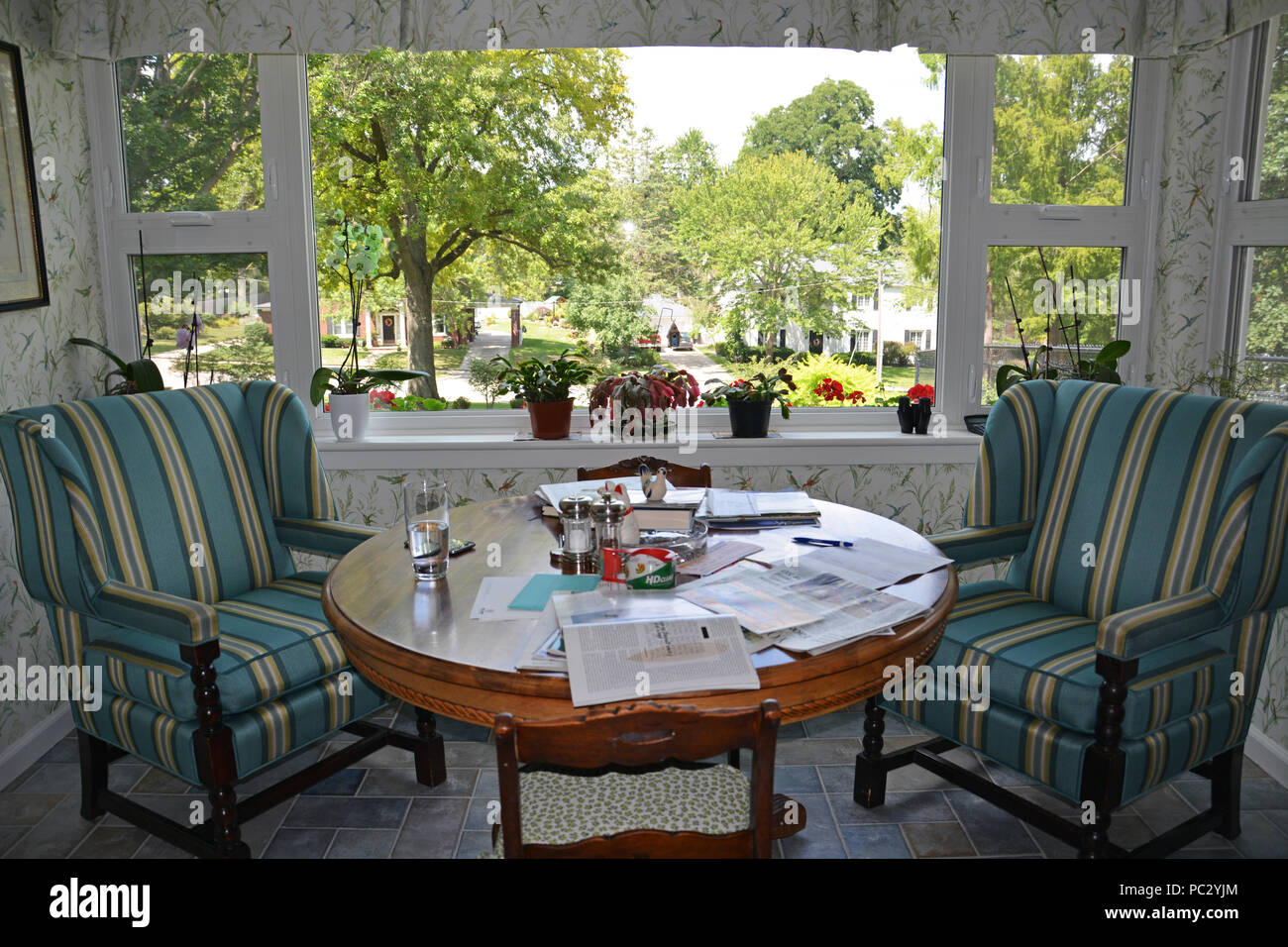 Un coin-repas en face de grandes fenêtres donnant sur le jardin avec deux chaises et table circulaire encombré d'éléments de la vie quotidienne Banque D'Images