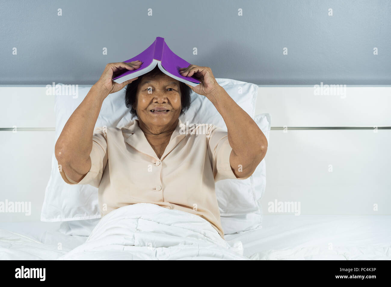 Heureux vieille femme au lit tenant un livre sur la tête. Banque D'Images