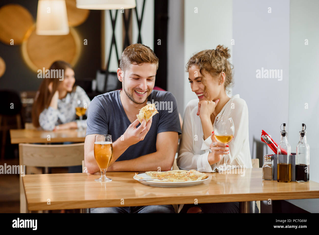 L'intérieur de la pizzeria. Sweet couple assis près de la table, de sourire et de poser. Belle femme avec des cheveux bouclés et handsome man eating pizza. Pizza et verres de vin et de la bière sur la table. Banque D'Images