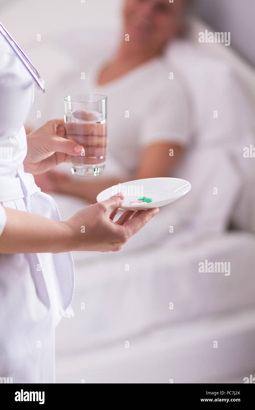 Image coupée de nurse holding midicine sur plaque. Femme médecin en blouse blanche remise pilules et verre d'eau au patient dans le lit. Banque D'Images