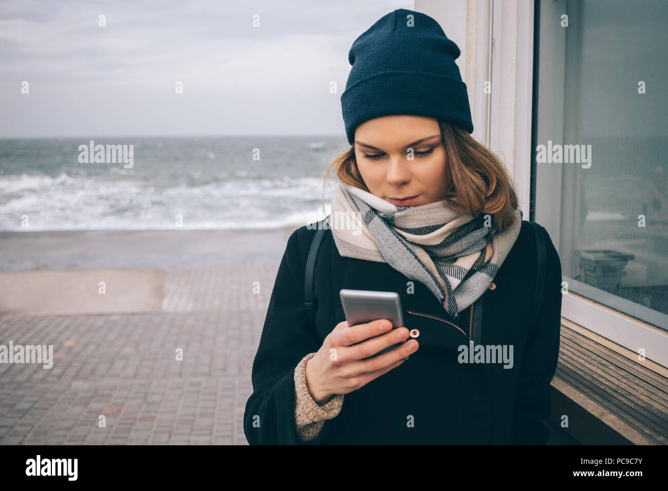 Jeune femme élégante en manteau, beanie bonnet et écharpe avec smart phone  près de la mer à la saison froide. Femme désirant en appareil mobile sur  journée d'hiver par th Photo Stock -