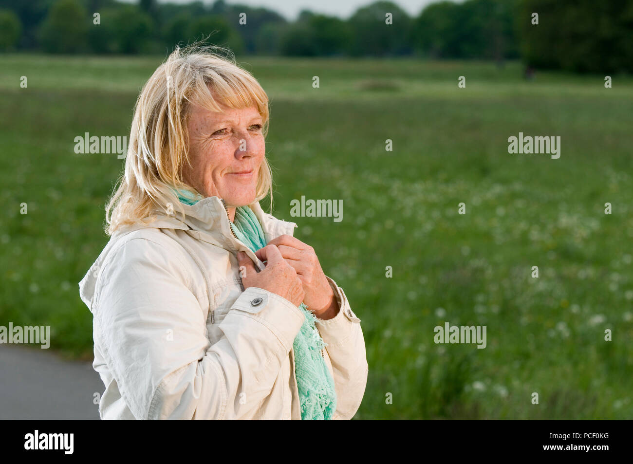 Les head and shoulders portrait of a middle-aged woman in front of green meadow et smiling sky à la recherche sur le côté. Les mains sur les revers Banque D'Images