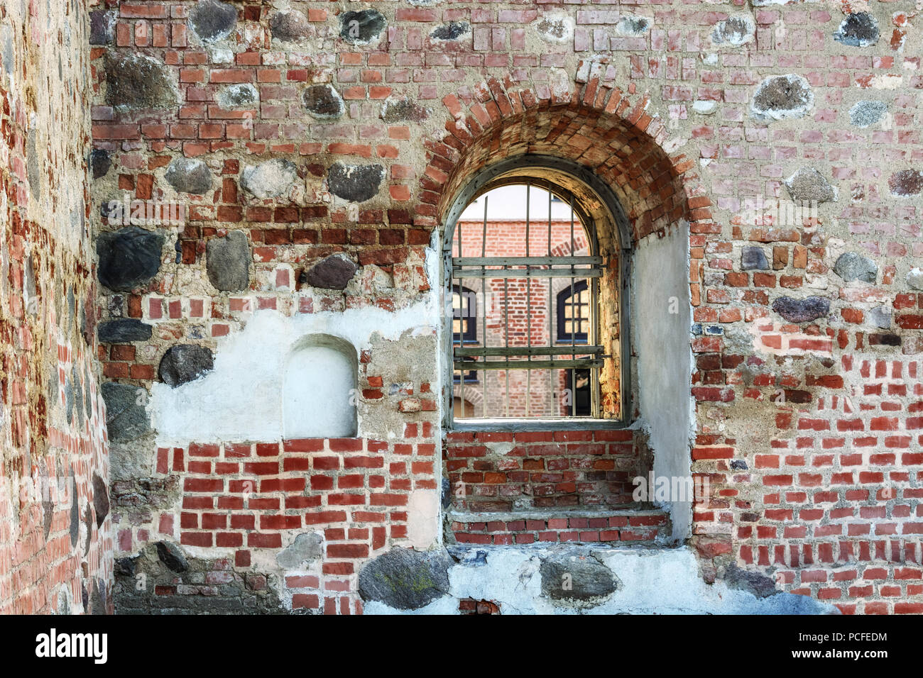 Une fenêtre avec un arc et fer à repasser grille dans un épais, vieille forteresse de brique mur à travers lequel la cour est visible. Banque D'Images