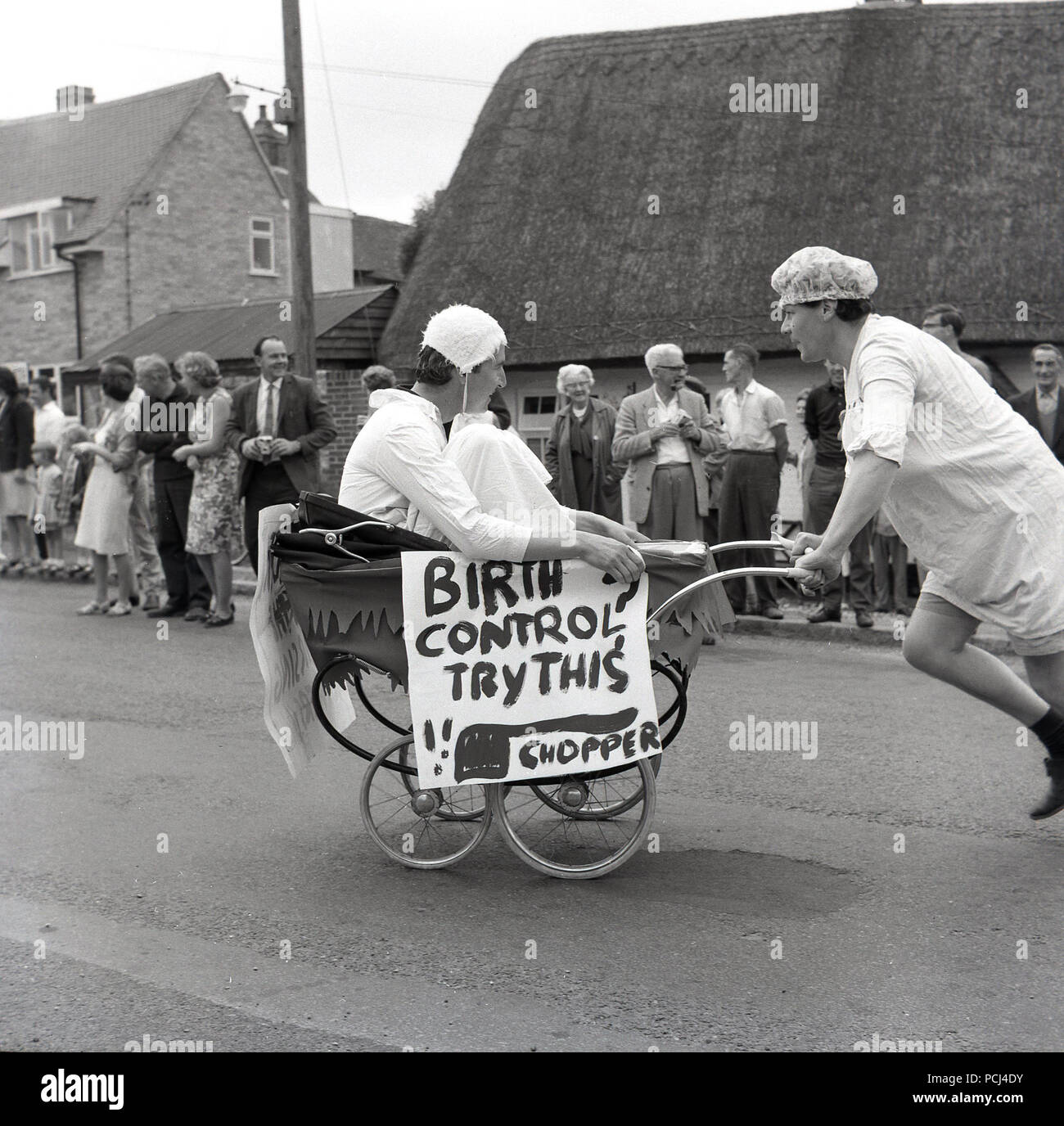 Années 1960, historique, village anglais, la pram race, deux concurrents  masculins dans les costumes, une séance dans un landau de l'époque, l'autre  poussant, passe le long d'une route pendant la course, observé