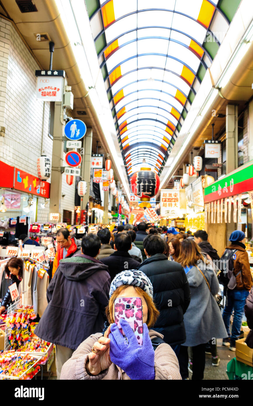 Kuromon Ichiba, Osaka's kitchen marché alimentaire. Woman with mobile phone selfies d'elle-même au marché avec des foules de gens derrière elle. Banque D'Images