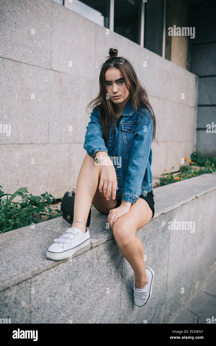 Teen girl posing in city portant tenue décontractée : short, veste bleu  jeans et baskets blanches. Portrait de femme à la mode avec bun hairstyle  assis nea Photo Stock - Alamy