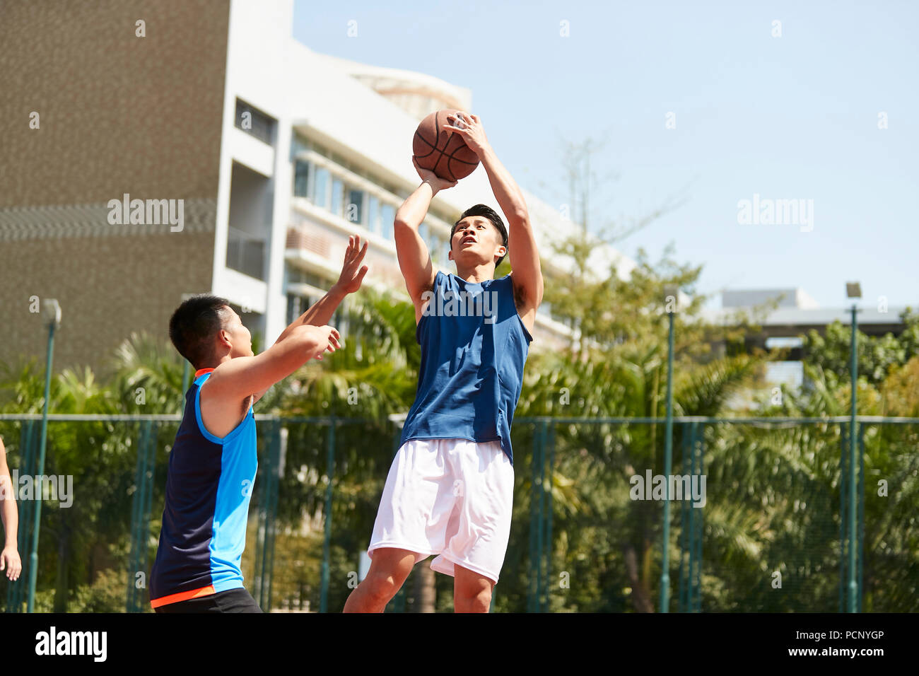 Young Asian basket-ball joueur qui place un tir alors que la défense adversaire jouer essayer de bloquer la balle. Banque D'Images