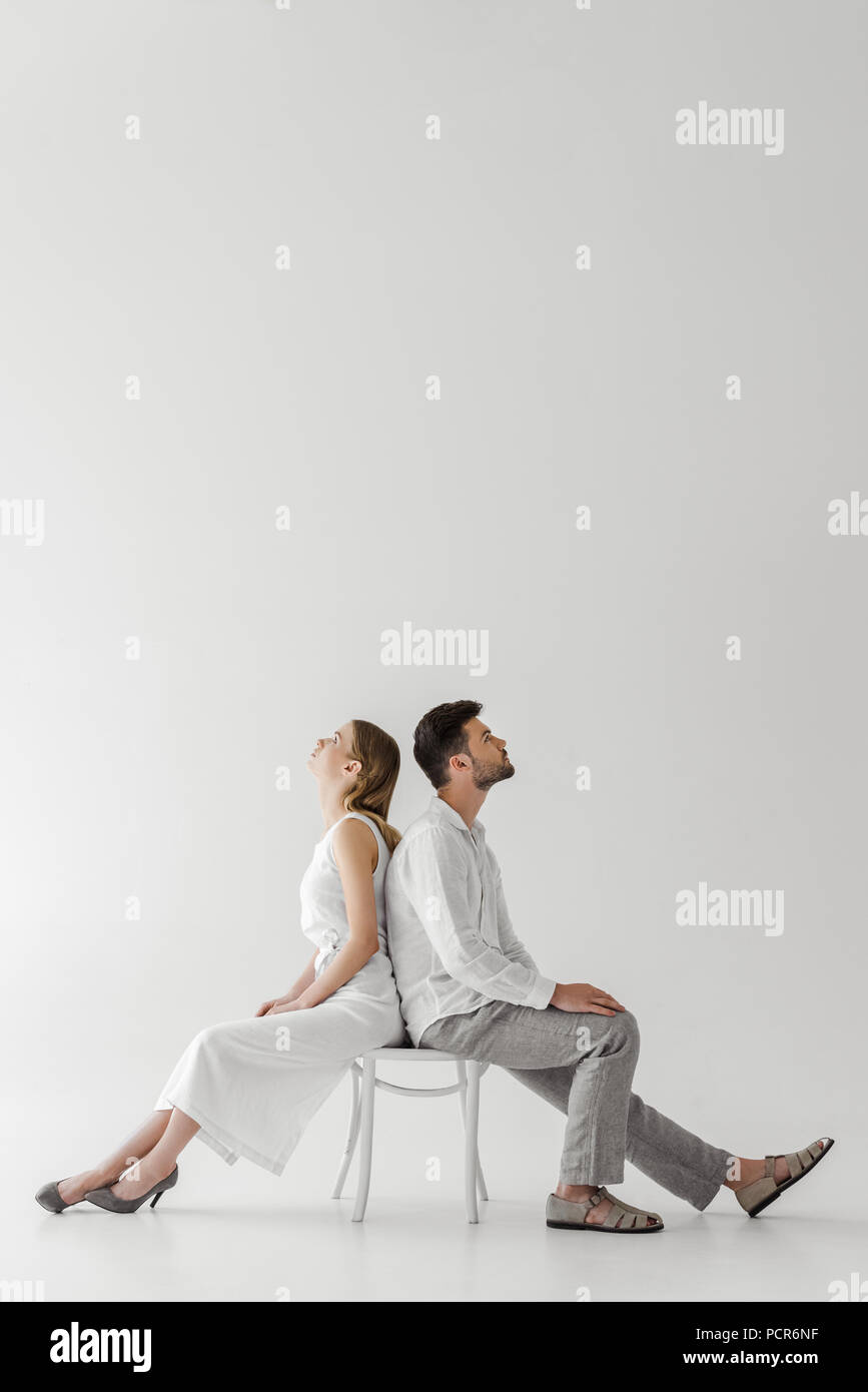 Vue latérale du jeune couple de modèles en lin vêtements assis dos à dos sur une chaise isolée sur fond gris Banque D'Images