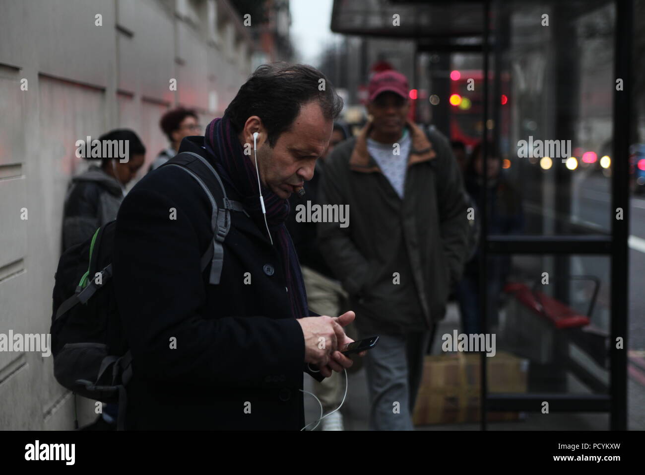 Soirée d'hiver voyageurs en attente sur un arrêt de bus sur Park Lane près de Marble Arch à Londres avec l'homme d'âge moyen à l'écoute via les câbles oreillettes smartphone Banque D'Images