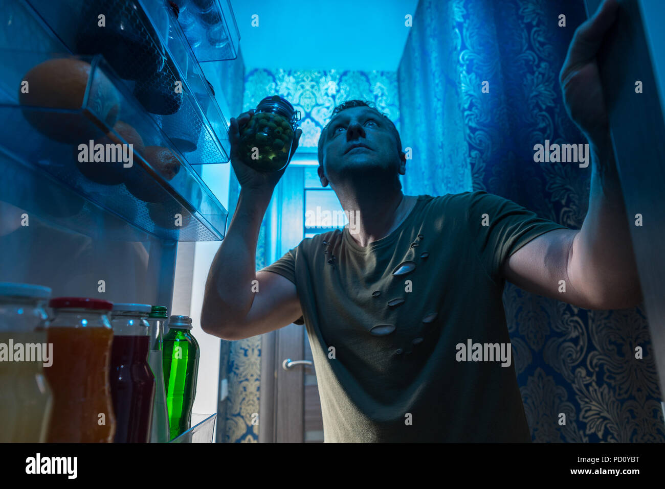 Homme recherche dans le réfrigérateur pour une collation de minuit debout dans l'obscurité de la porte ouverte la tenue d'un bocal de cornichons et à la recherche de nourriture sur l'étagère Banque D'Images