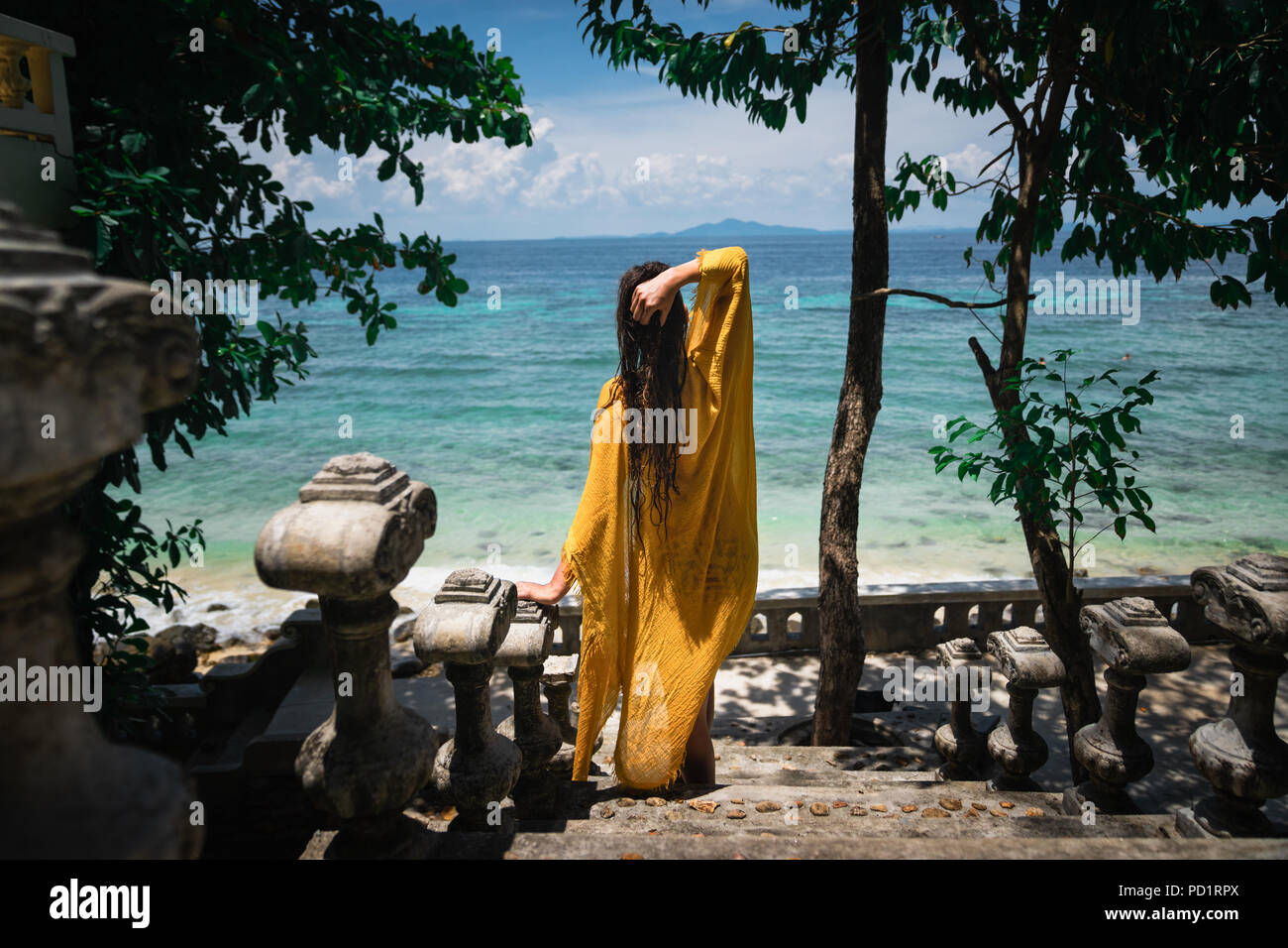 Jeune belle femme bronzée dans une robe jaune sur la plage en face de l'océan sur un escalier en pierre Banque D'Images