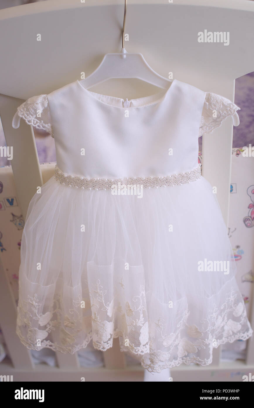 Adorable bébé fille robe blanche, accrochée à un cintre petite, vertical, vue frontale, contre lit bébé blanc Banque D'Images