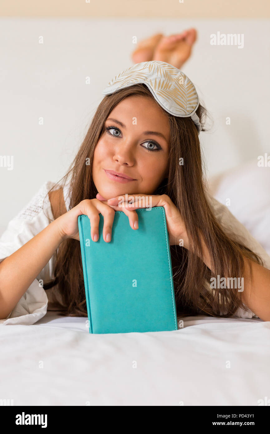 Jeune femme en pyjama et masque pour les yeux en posant la peau parfaite à jeter au lit après service et de tenir son livre de calendrier. Belle fille sur whit Banque D'Images