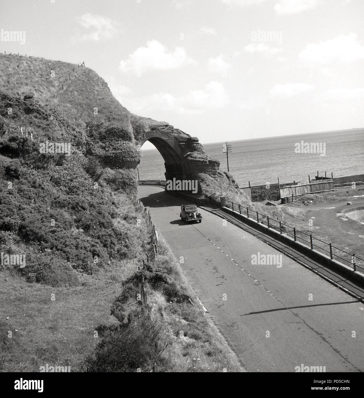 Années 1950, tableau historique par j Allan l'argent comptant, une voiture de l'époque voyageant sur un quartier calme, à vide, sur la spectaculaire route côtière causeway Antrim, Irlande du Nord, Royaume-Uni. Banque D'Images