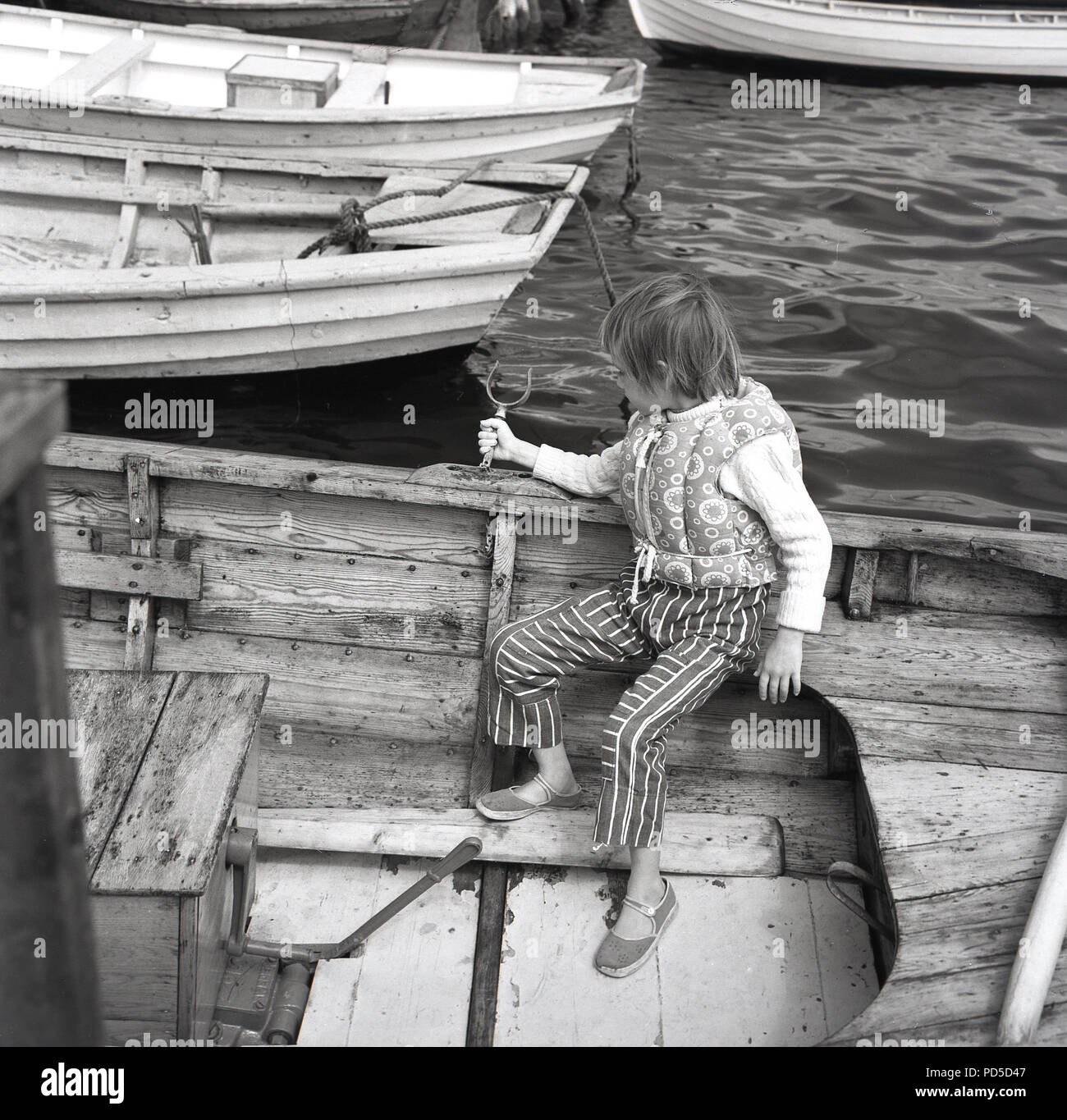 Années 1960, jeune fille stting sur son propre avec son gilet sans manches sur à l'arrière d'un bateau à rames en bois dans un port, England, UK. Banque D'Images