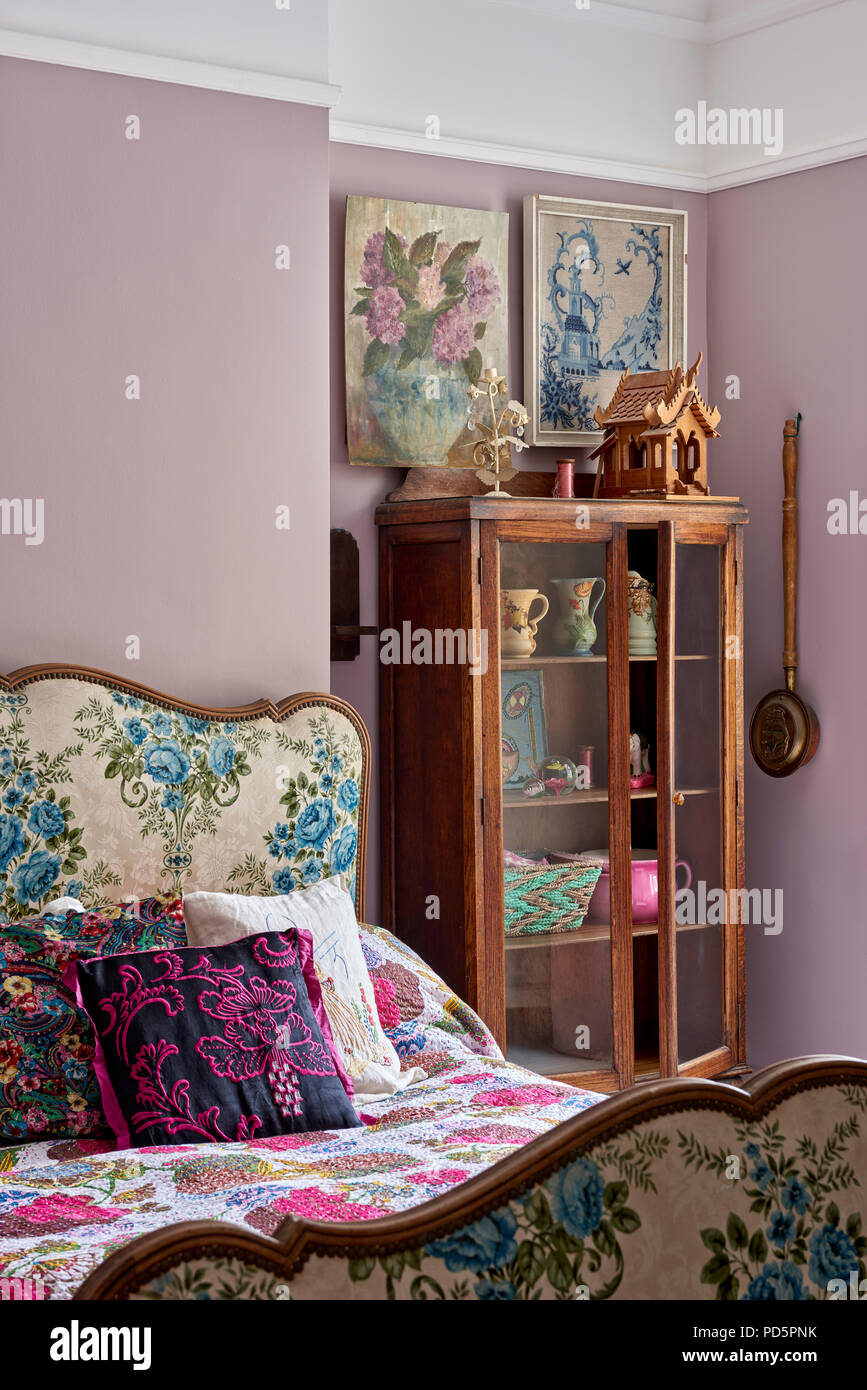 Lit français rembourrés en tissu floral vintage dans la chambre peinte de couleur violet poussiéreux. La façade de verre display cabinet est Edwardian. Banque D'Images