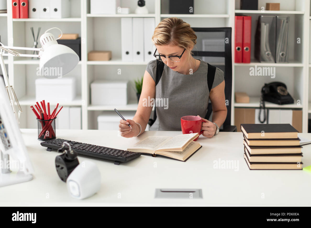 Une jeune fille est assise à une table dans le bureau, tenant un crayon et  une tasse rouge dans sa main. Avant de la jeune fille se trouve un livre  ouvert Photo