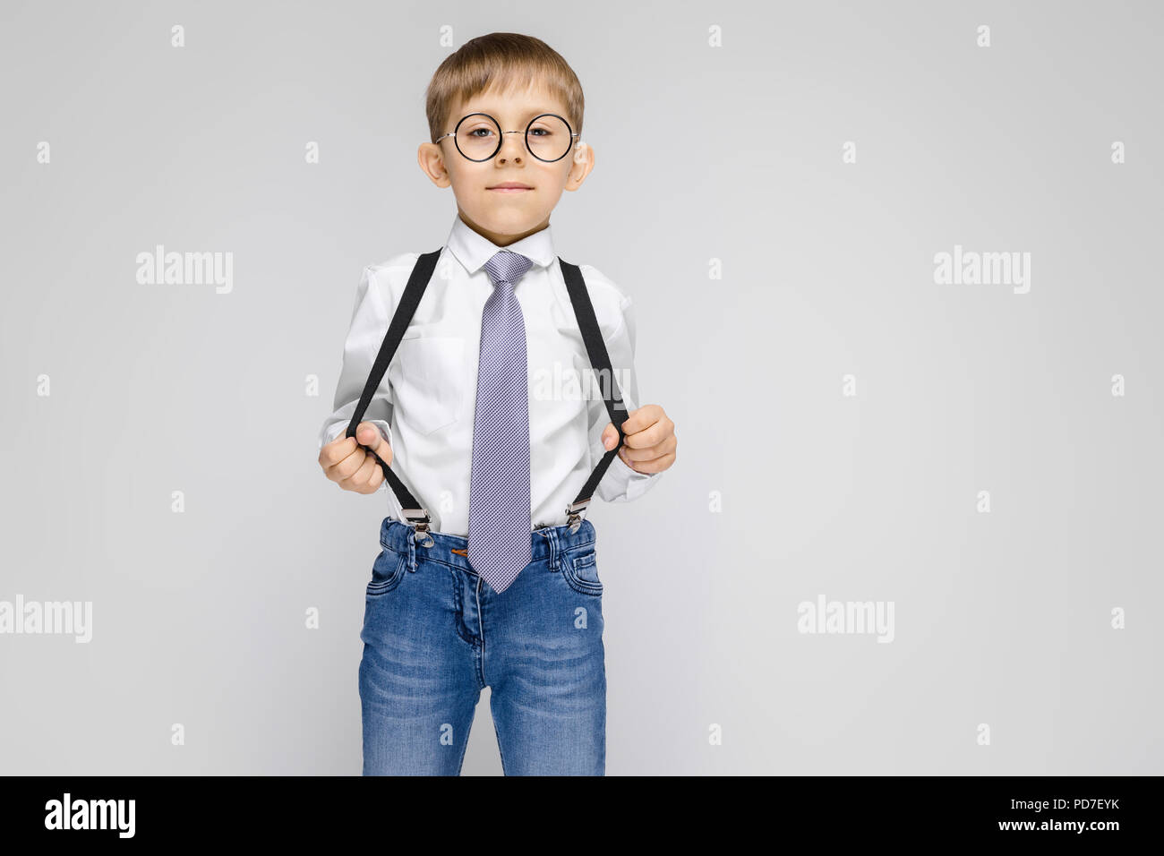 Un charmant garçon dans une chemise blanche, bretelles, une cravate et des  jeans se dresse sur un fond gris. Le Garçon à lunettes tiré les bretelles  Photo Stock - Alamy