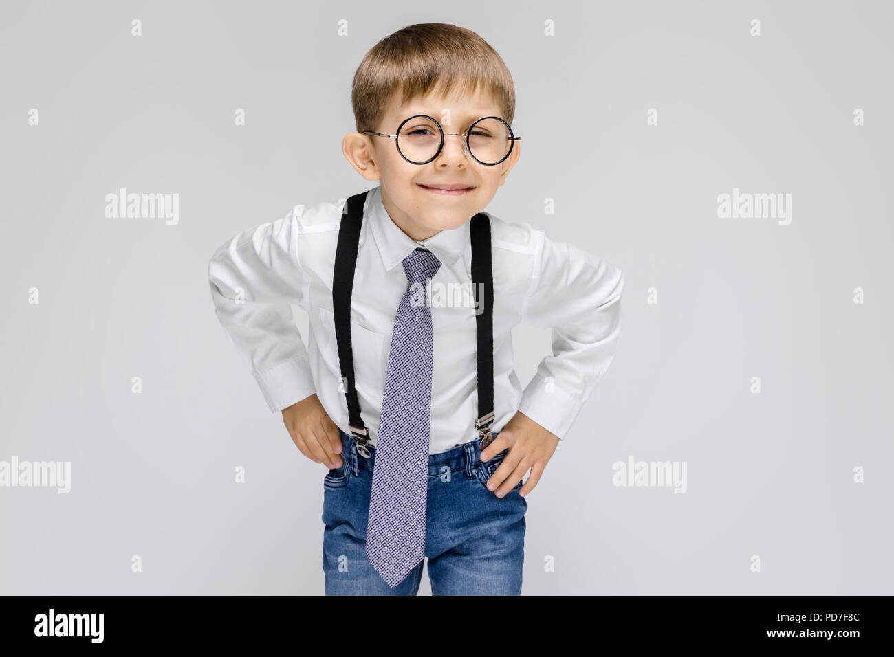 Un charmant garçon dans une chemise blanche, bretelles, une cravate et des  jeans se dresse sur un fond gris. le garçon avec des lunettes à l'écoute de  ce qu'il est dit Photo
