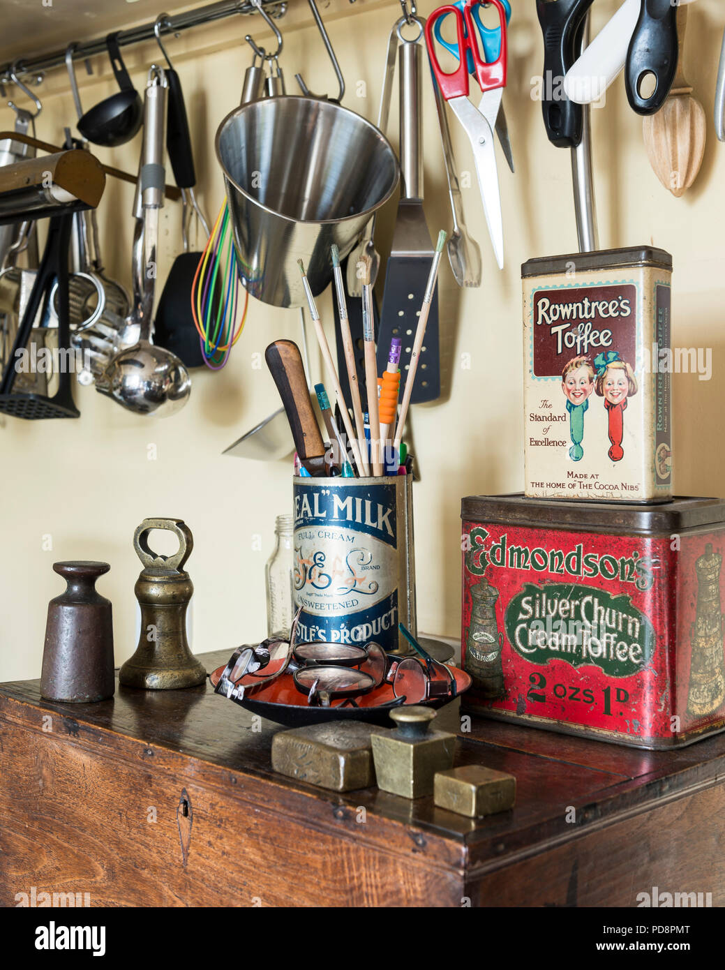 Les poids en laiton et vintage sweet tins s'asseoir sur une vieille boîte de chêne. Au-dessus de divers ustensiles de cuisine se suspendre à un mur rack Banque D'Images