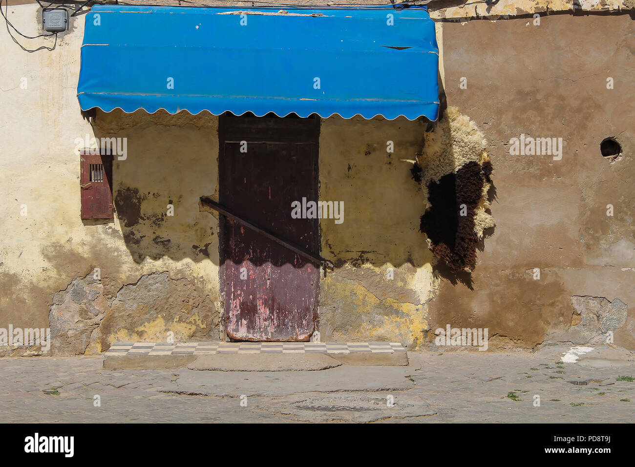 Vue d'une rue sur une porte fermée d'une boutique dans une maison ancienne. Marquise de l'ombre bleue pour certains. La peau de mouton accroché près de la porte. El Jadida, Maroc. Banque D'Images