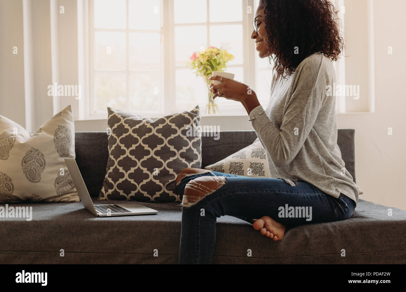 Femme souriante dans un Jean déchiré à la mode tenant une tasse à café tout en travaillant sur un ordinateur portable à la maison. femme d'affaires assise sur le canapé à la maison et au travail Banque D'Images