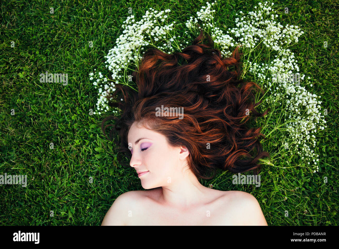 Closeup portrait de jeune fille belle femme aux cheveux brun rouge couché sur l'herbe avec de petites fleurs blanches autour de la tête. Vue de dessus haut overhe Banque D'Images