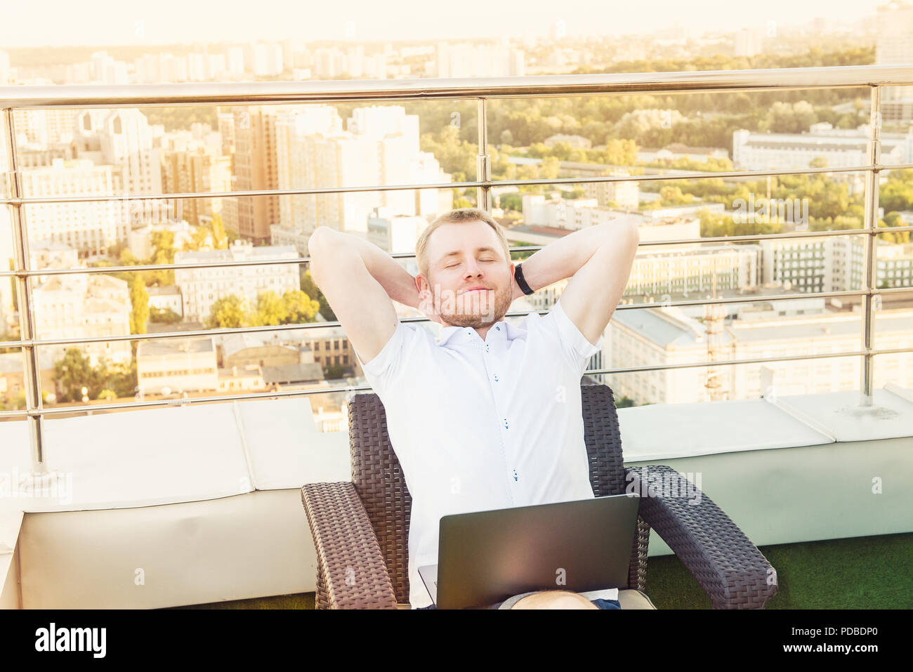 Satisfaits de travailler. Cheerful young man holding hands behind head et de garder les yeux fermés, assis sur la terrasse avec vue sur le paysage urbain Ba Banque D'Images
