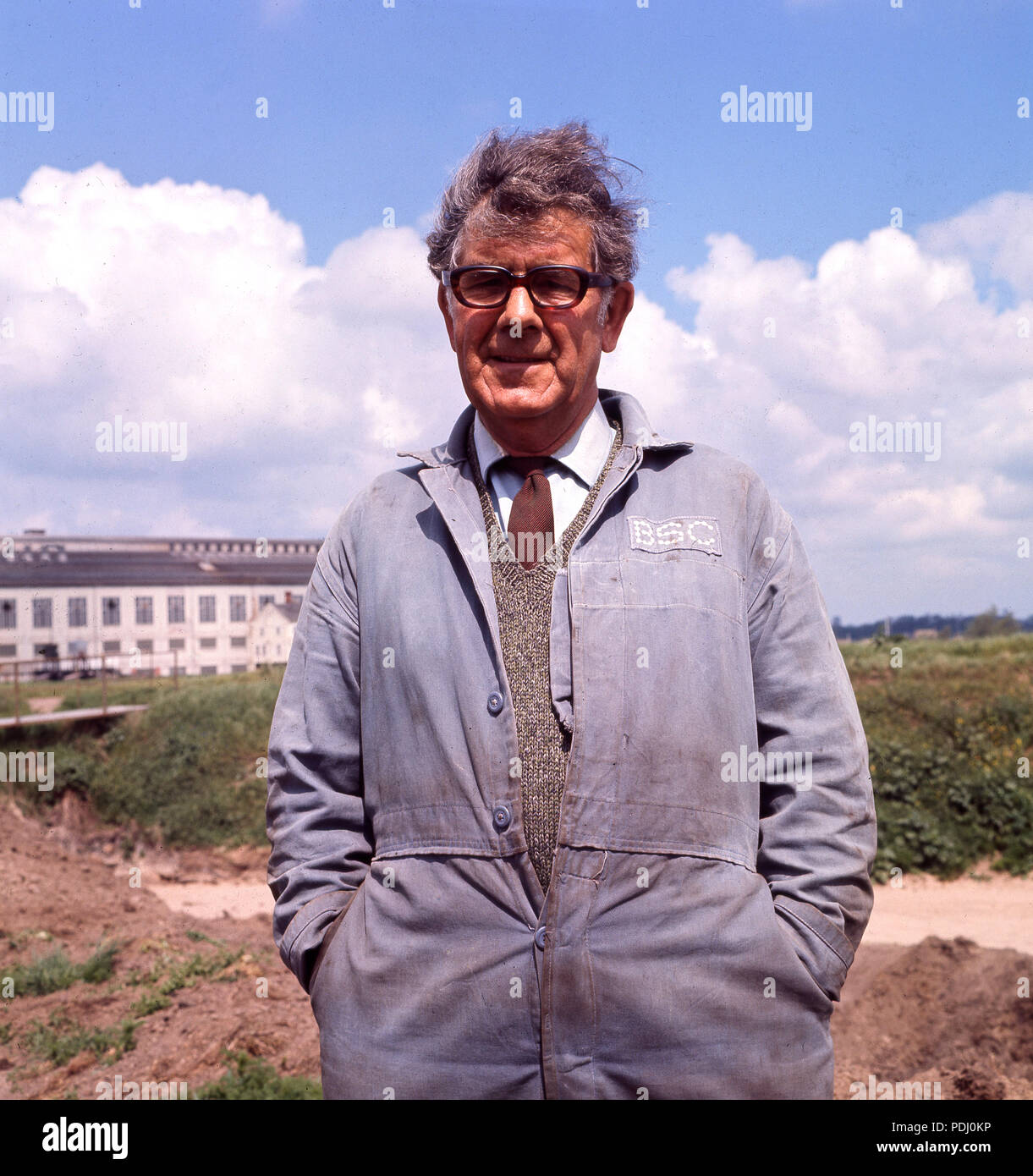 1960, l'homme debout à l'extérieur dans une chemise et une cravate et des vêtements de travail, le bleu/gris bleu de travail de la British Steel Corporation (BSC), Angleterre, Royaume-Uni. Banque D'Images