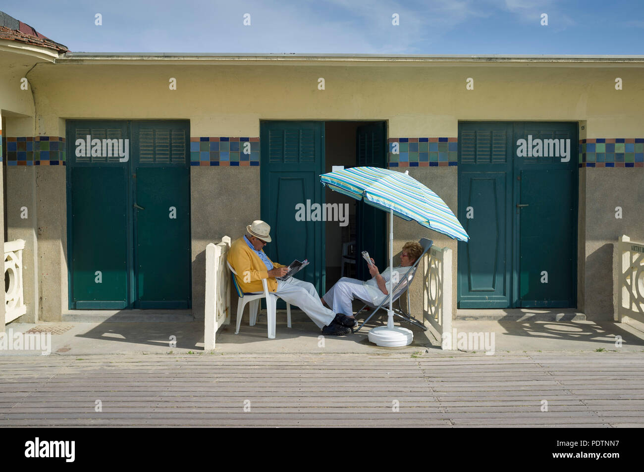 Un couple mature lire en dehors de leur cabine de plage sous un parasol sur la promenade des Planches de Deauville, Normandie, France Banque D'Images