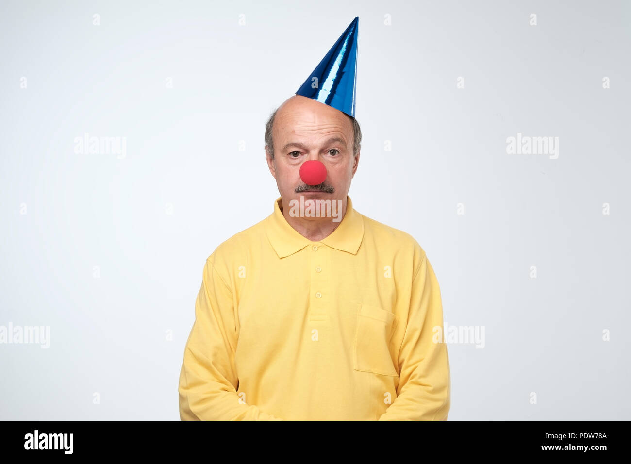 Triste anniversaire gars en t-shirt jaune et rouge nez de clown debout sur fond blanc. Tout le monde a oublié son parti. Être seul concept Banque D'Images