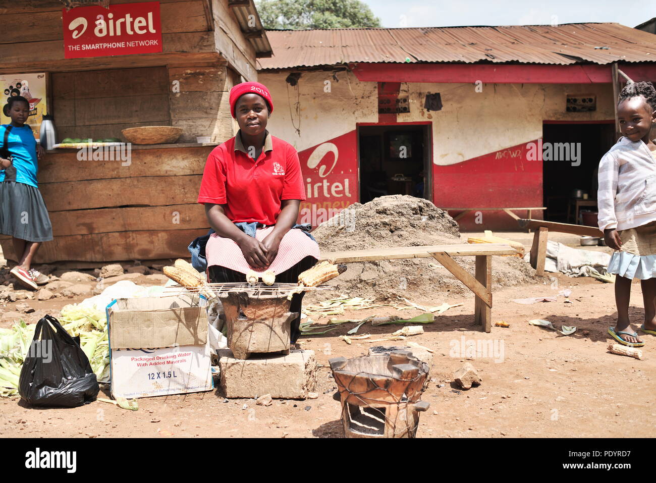 Jeune mère ougandaise est assis sur un siège en bois dans une petite ville, de vendre le maïs et autres cultures de gagner leur vie. Banque D'Images