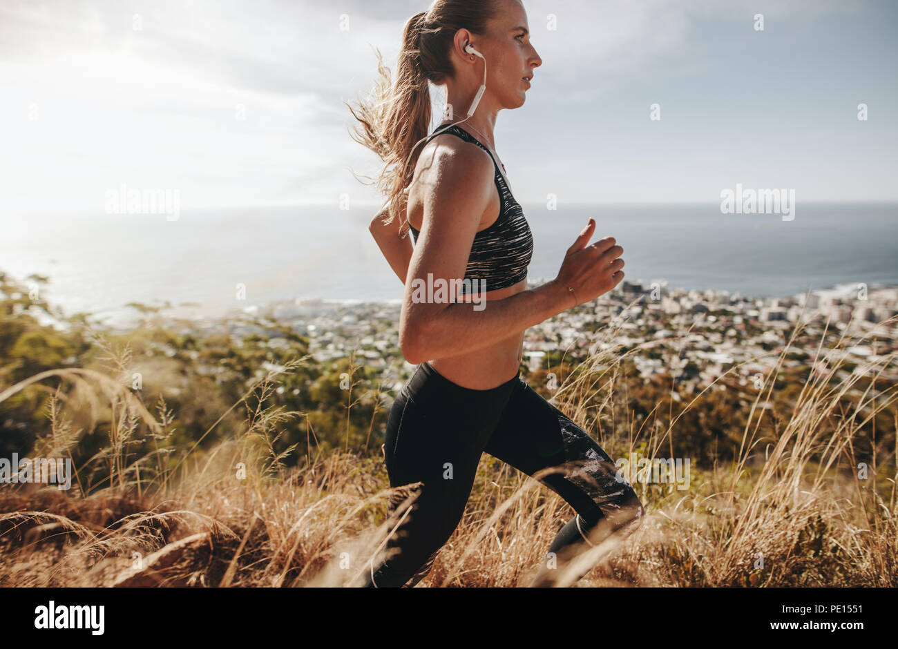 Vue latérale du fit young woman wearing earphones traversant le sentier de montagne. Coureuse sur formation sentier dans la colline. Banque D'Images