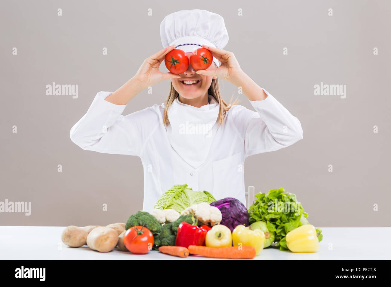 Cheerful female chef est couvrant ses yeux avec de la tomate alors qu'il était assis à la table avec des tas de légumes. Banque D'Images