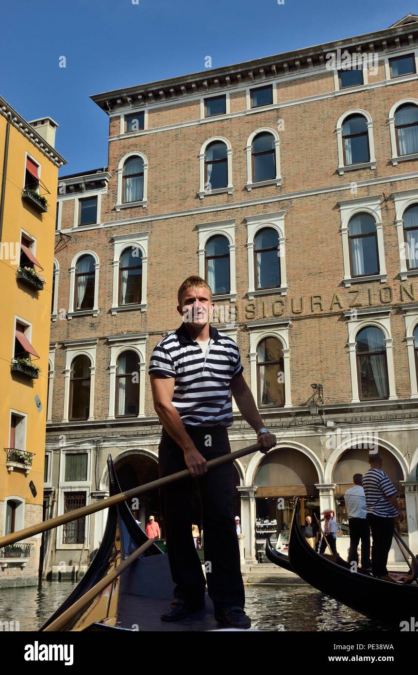 Une vue en contre-angle d'un gondolier, portant l'emblématique T-shirt noir et blanc et rasant sa gondole, contre des bâtiments historiques, Venise, Italie, Europe Banque D'Images