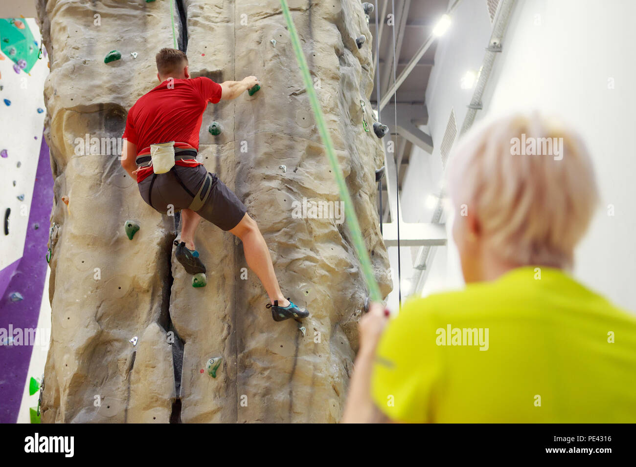 Photo de l'arrière de sportsman grimper sur un mur d'escalade et d'entraîneur féminin tenant la corde de sécurité Banque D'Images