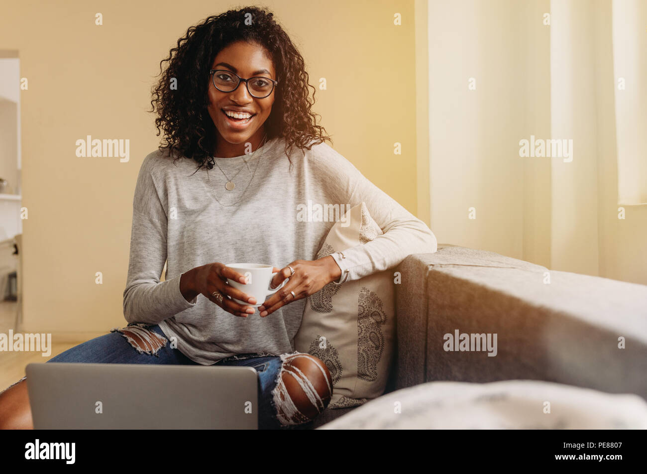 Smiling woman mode dans le jeans déchirés tenant une tasse de café tout en travaillant sur un ordinateur portable à la maison. Businesswoman sitting on sofa at home et worki Banque D'Images