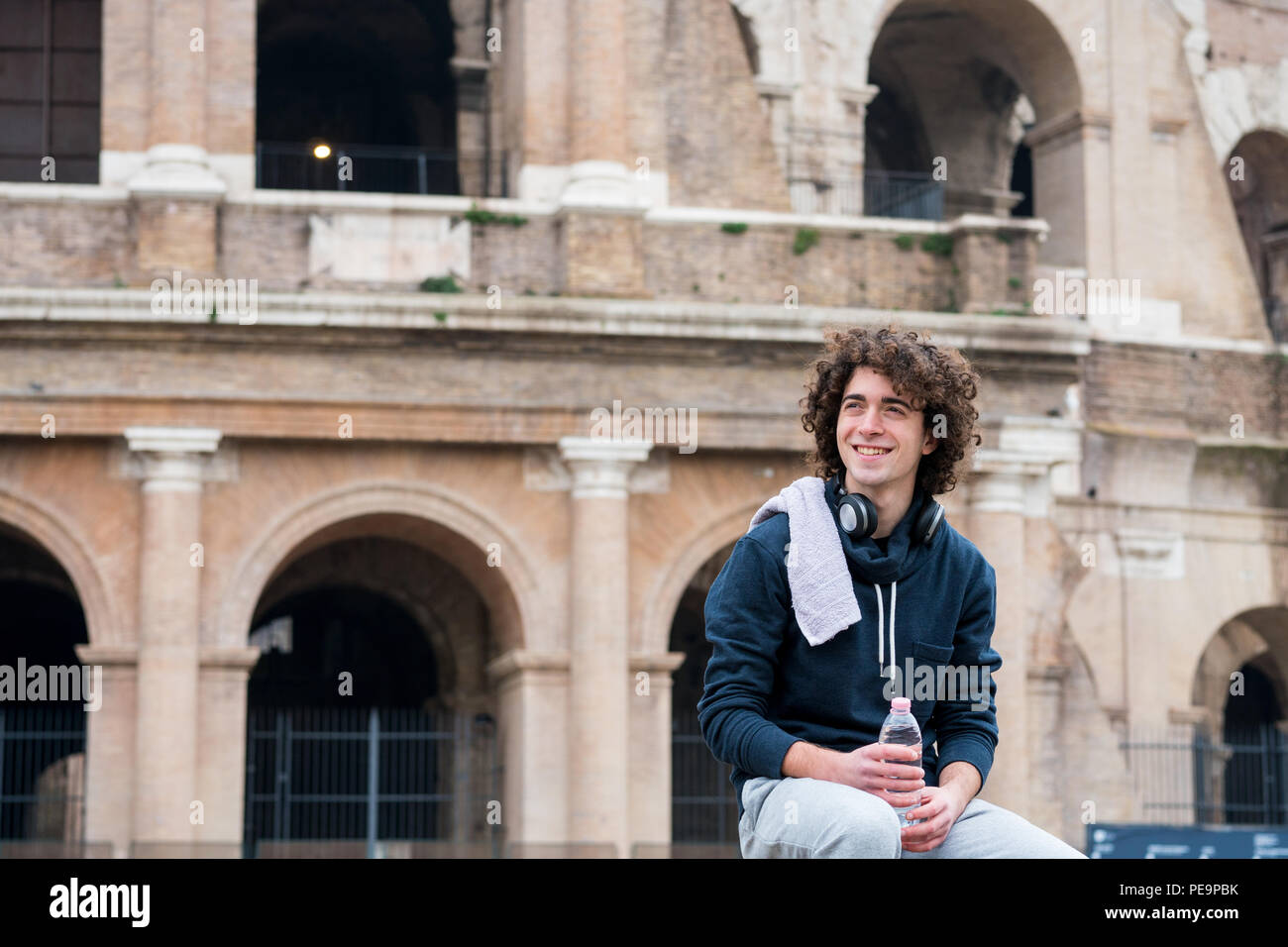 Beau jeune sportsman holding water bottle et relaxant après une formation avec une serviette autour de son cou en avant du Colisée à Rome Banque D'Images