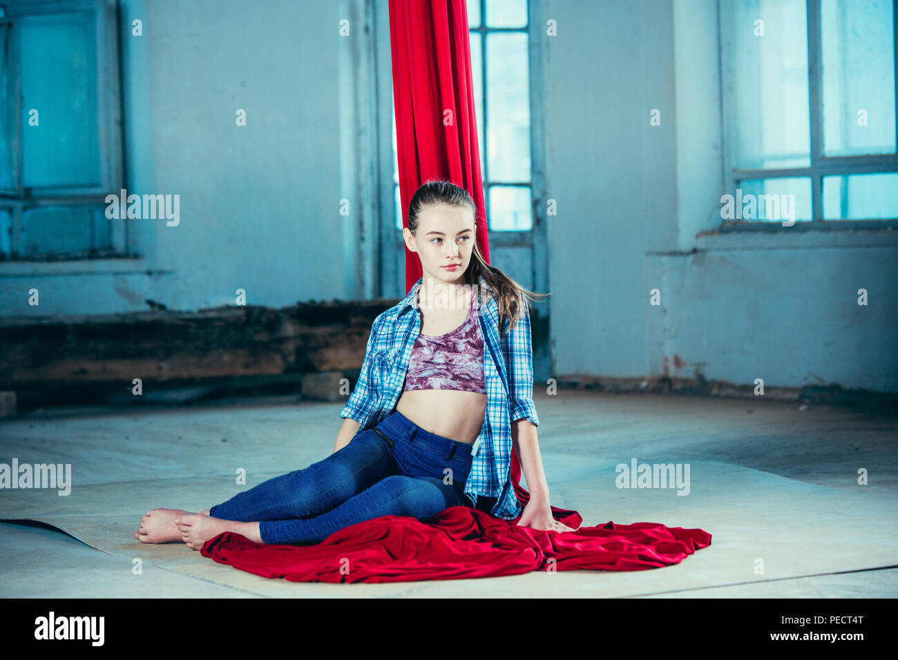 Gymnaste gracieuse se reposant après l'exécution de l'exercice aérien avec des étoffes rouges sur fond bleu ancien loft. Young teen caucasian fit girl. Le cirque, acrobatie, Acrobat, interprète, sport, fitness, gymnastique concept Banque D'Images