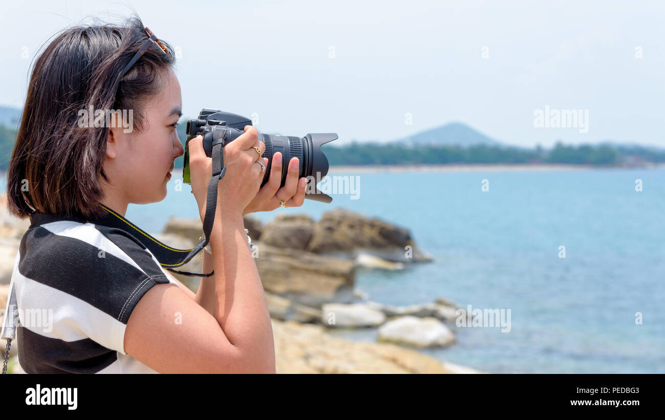 Jeune femme était heureux à la photographie avec l'appareil photo reflex numérique sur le rocher près de la mer sous le ciel d'été à l'île de Koh Samui, Surat Thani province, Thaïlande Banque D'Images