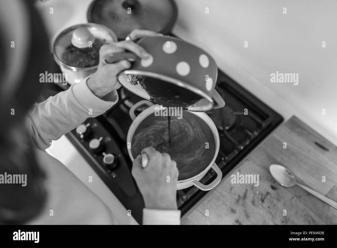 Image en niveaux de gris de la femme la cuisson verser la crème au chocolat d'un pot dans un deuxième pot sur la table de cuisson d'une cuisinière dans la cuisine, vue du dessus. Banque D'Images