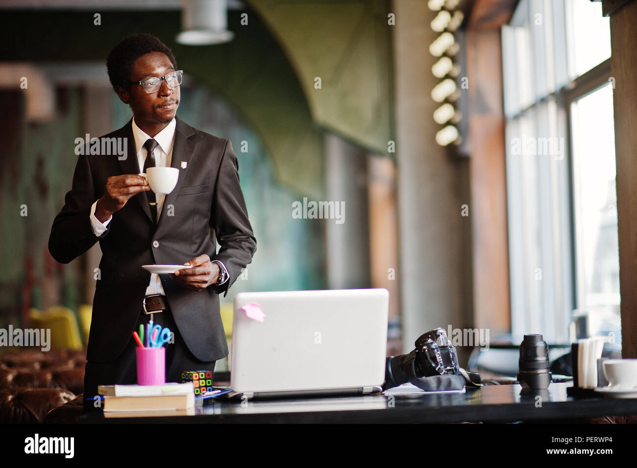 African American man photographe paparazzi de l'usure sur un costume noir et de lunettes au bureau avec ordinateur portable, de boire un café le matin avant le jour de travail. Banque D'Images