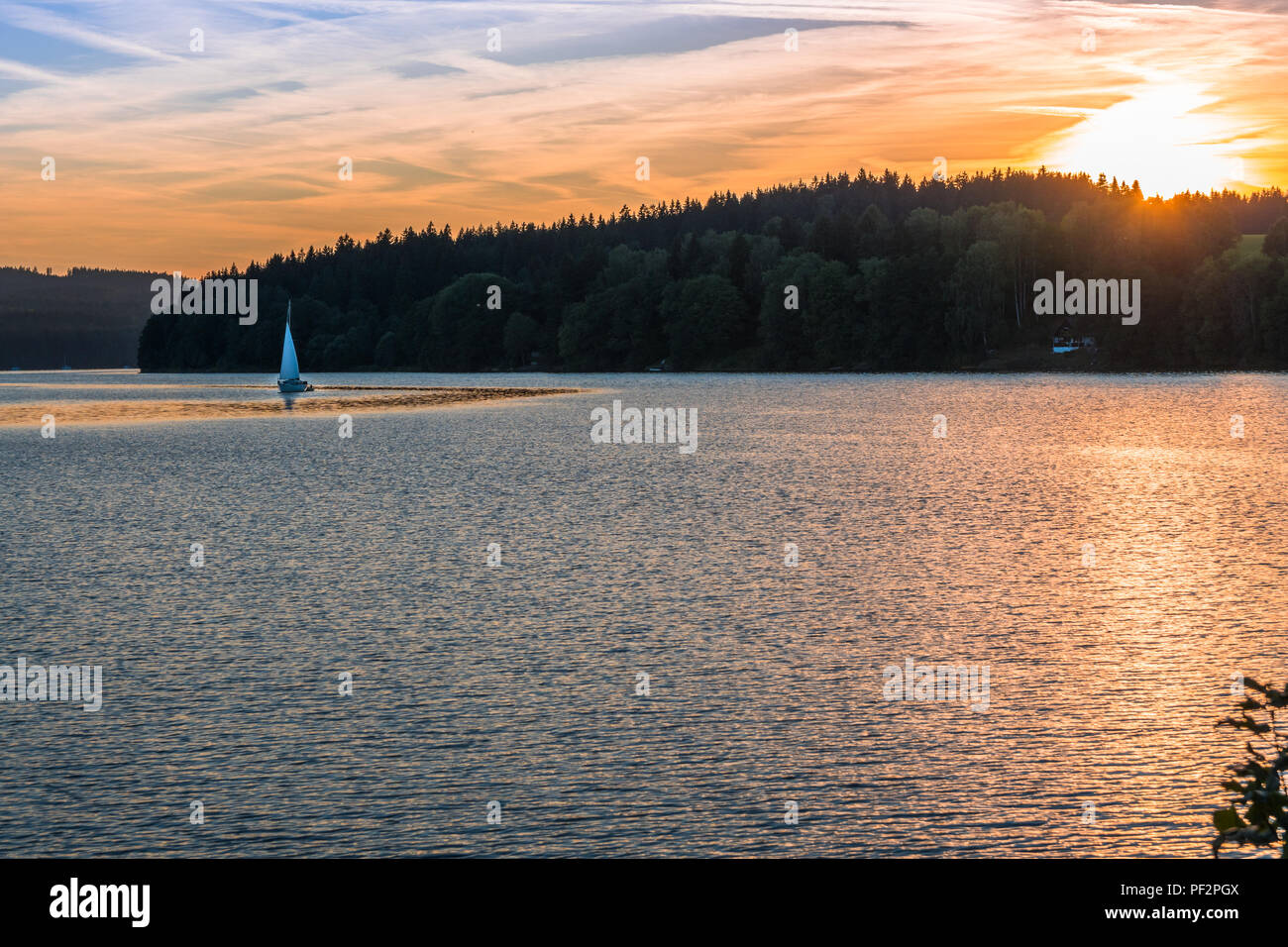 Avec un romantique coucher de soleil orange derrière des arbres au-dessus d'un grand lac avec de petites vagues avec le petit yacht de voile sur un plan d'eau. Bon été golden hour sho Banque D'Images