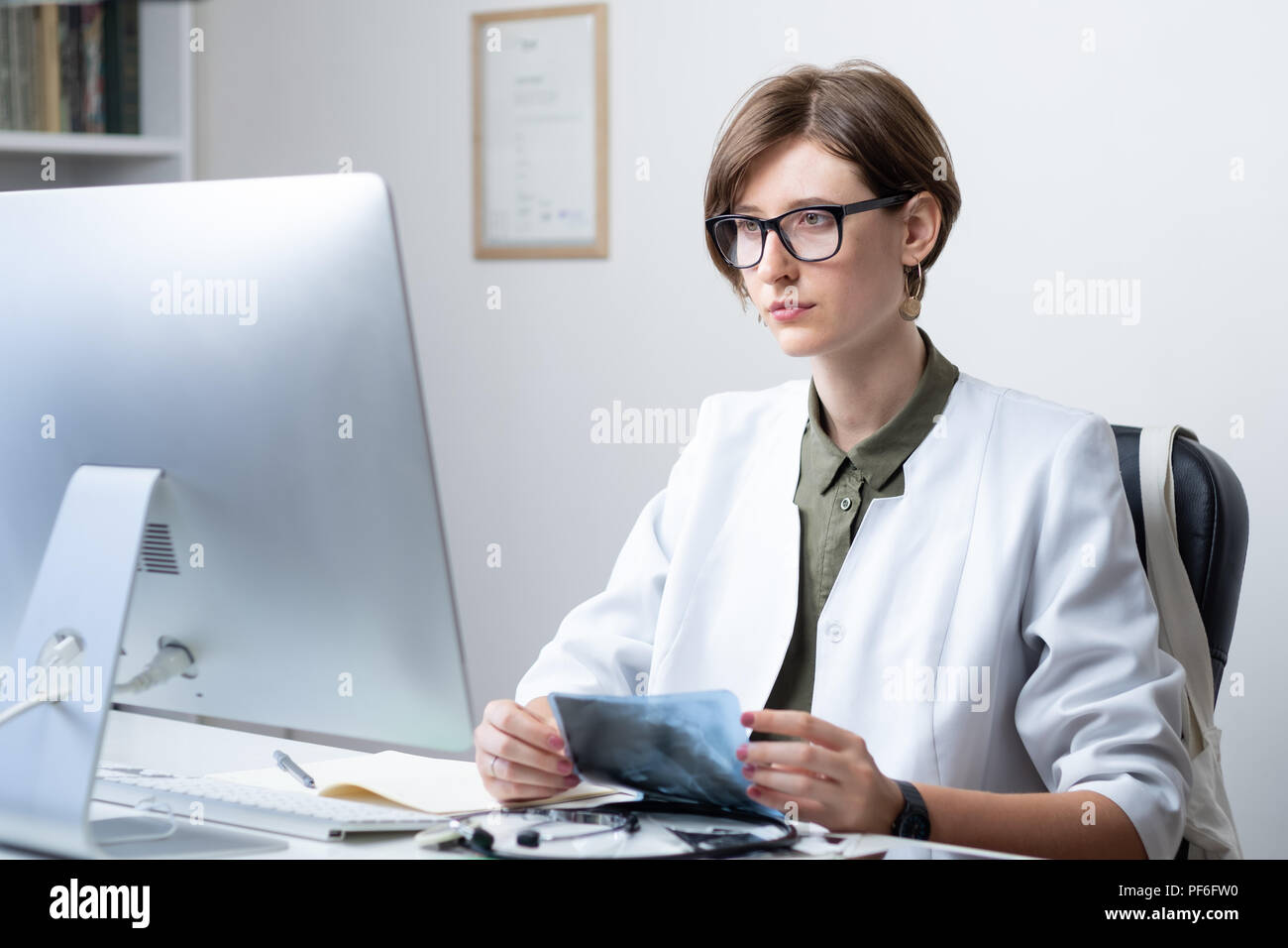 Femme médecin praticien au bureau moderne. Jeune médecin examining x-ray sur le lieu de travail devant un ordinateur de bureau Banque D'Images