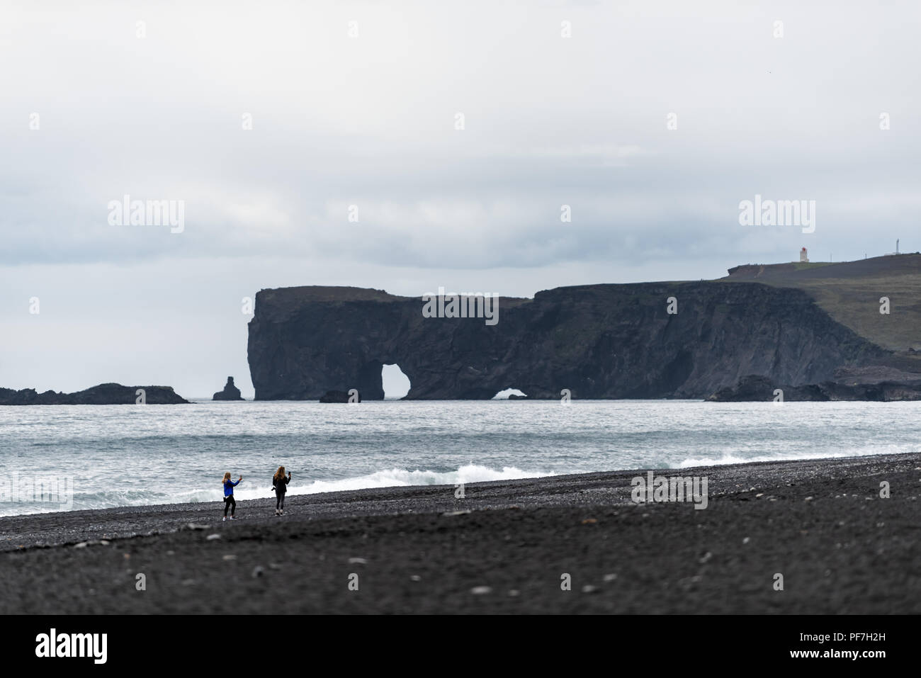Islande Vik, deux jeunes femmes touristes sur black sand beach, ocean horizon, rock formation arch à Dyrholaey, Kirkjufjara photographier le phare sur so Banque D'Images