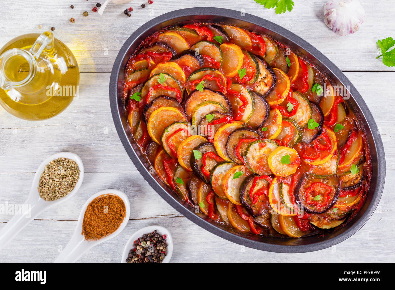 Ratatouille en couches dans un plat allant au four, des tranches de  courgette, poivron rouge, piment, aubergines, courgettes jaunes, l'huile  d'olive, le persil et l'ail, sur fond blanc, ba Photo Stock -