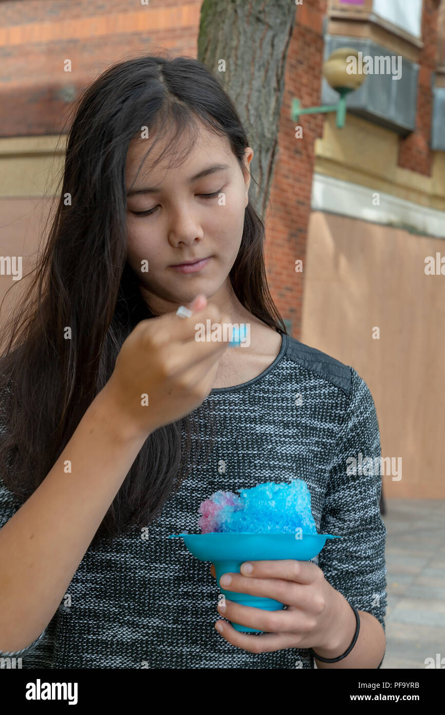 Français/Thaï adolescente avec de longs cheveux noirs mange une caisse noire bleue dans le centre-ville de Warrington Banque D'Images