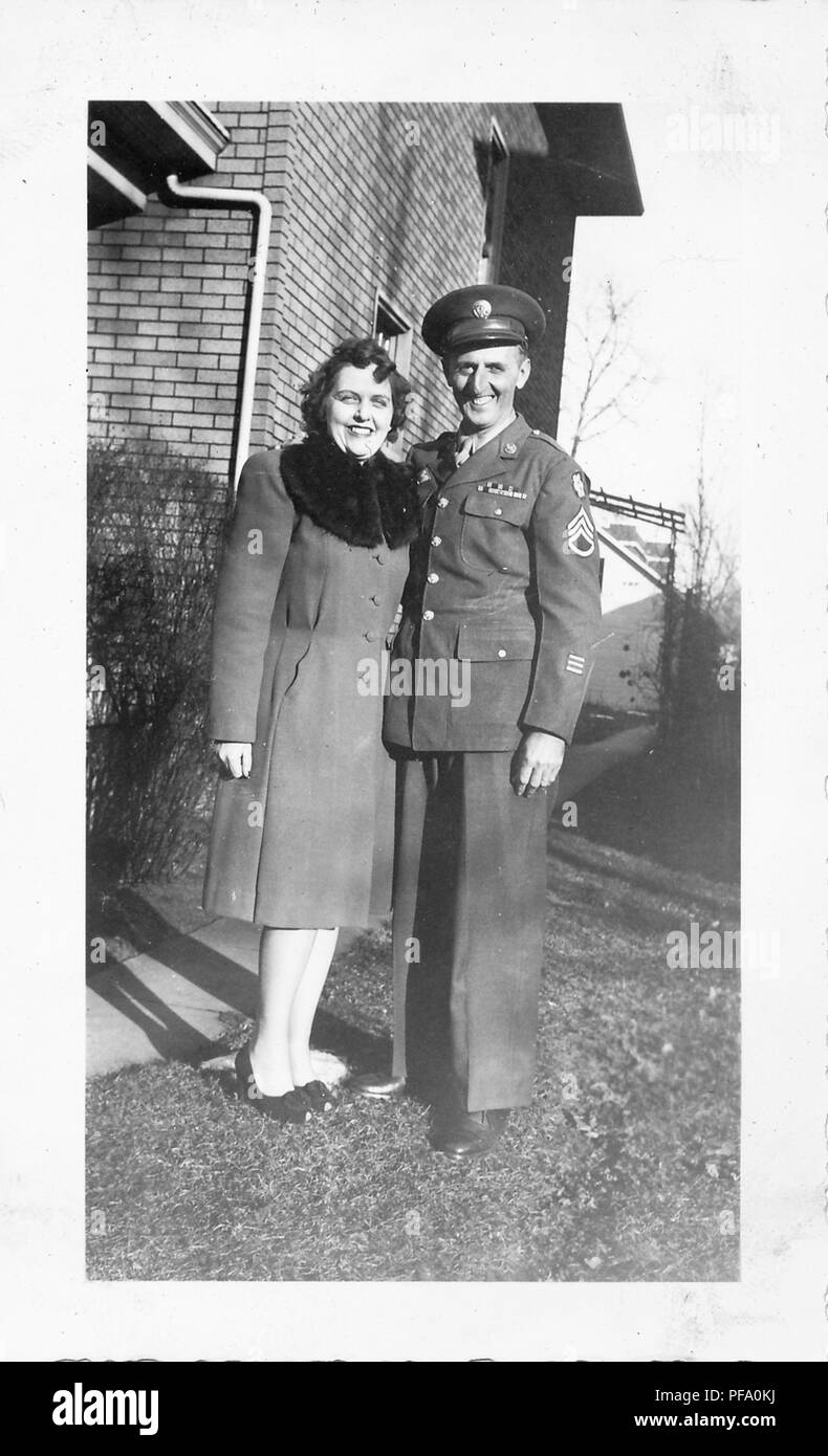 Photographie noir et blanc, tourné à partir d'un angle faible, montrant un couple souriant, à la fois debout dans toute la longueur, et face à la caméra, l'homme portant un uniforme militaire et la cap, le dark-haired woman wearing bow-front, peep-orteils, talons et d'un manteau d'hiver longueur genou avec un col de fourrure sombre, avec un bâtiment en brique visible à l'arrière-plan, probablement photographié dans l'Ohio pendant la Seconde Guerre mondiale, 1950. () Banque D'Images